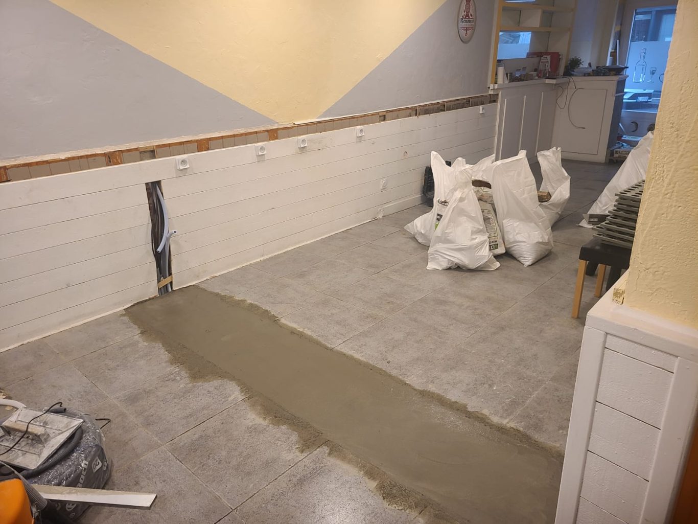 RÉNOVATION DES CUISINES D'UN RESTAURANT : ÉLECTRICITÉ, PLOMBERIE ET INSTALLATION DE MATÉRIEL DE CUISINE Salle en rénovation avec sol en béton frais et sacs de matériaux entassés.