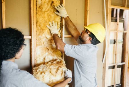 LES DIFFERENTS TYPES D'ISOLATION : COMMENT CHOISIR POUR VOTRE HABITAT Deux ouvriers installent de l'isolant dans un mur, l'un portant un casque de sécurité.